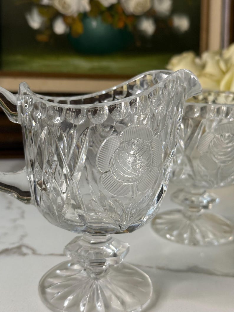 May include: Clear glass creamer with a rose design. The creamer has a handle and a spout. The base is a pedestal design. The creamer is part of a set. The glass has a diamond pattern.