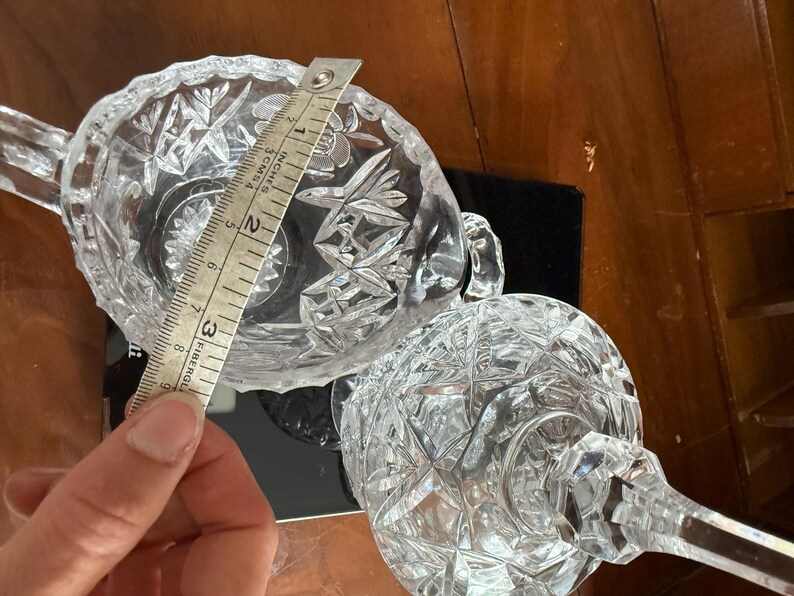 May include: Clear crystal sugar bowl and a stemmed glass with a diamond pattern. The sugar bowl is approximately 3 inches (7.6 cm) in diameter. The objects are on a dark surface.
