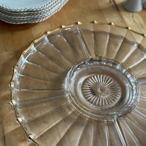 May include: A clear glass serving platter with a gold rim and a radial design. The platter has a central recessed area and a scalloped edge. A stack of white plates with a decorative border and a floral design are visible in the background.