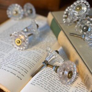 May include: Clear glass cabinet knobs with a faceted design and brass-colored hardware. Several knobs are arranged on an open book with aged pages. The knobs are a decorative element for furniture.