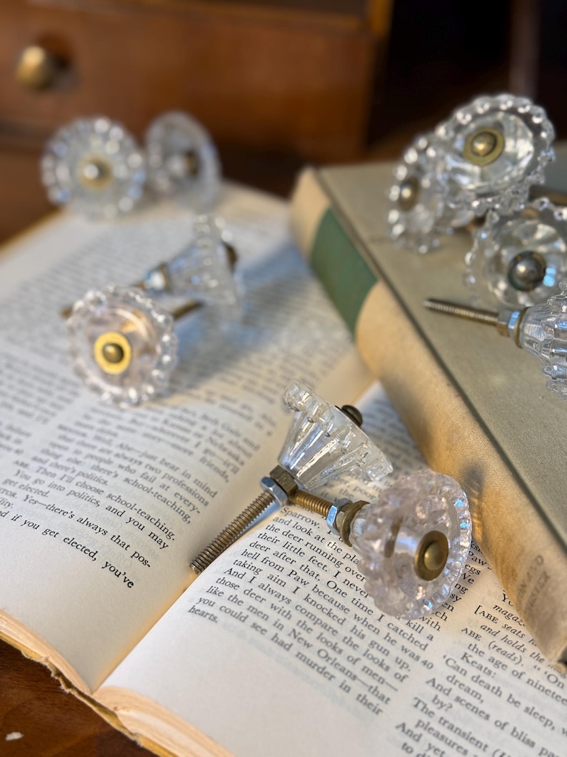 May include: Clear glass cabinet knobs with a faceted design and brass-colored hardware. Several knobs are arranged on an open book with aged pages. The knobs are a decorative element for furniture.