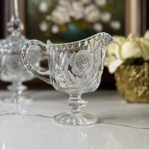 May include: A clear, vintage crystal creamer with a rose design. The creamer features a handle and a pedestal base. The crystal is cut with a diamond pattern. A matching sugar bowl is slightly out of focus in the background.
