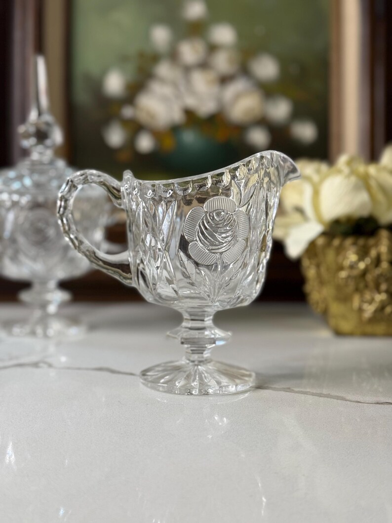 May include: A clear, vintage crystal creamer with a rose design. The creamer features a handle and a pedestal base. The crystal is cut with a diamond pattern. A matching sugar bowl is slightly out of focus in the background.