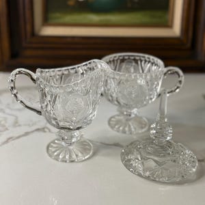 May include: Clear glass creamer and sugar bowl set with a rose pattern. The creamer has a handle, and the sugar bowl has a lid. The set is on a white surface, with a painting in the background.