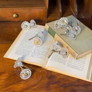 May include: Clear glass cabinet knobs with faceted edges and brass hardware are arranged on an open book and a closed book. The knobs have a vintage aesthetic and are displayed on a wooden surface.