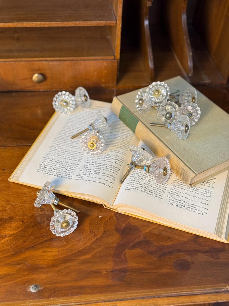 May include: Clear glass cabinet knobs with faceted edges and brass hardware are arranged on an open book and a closed book. The knobs have a vintage aesthetic and are displayed on a wooden surface.