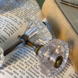 May include: Clear glass cabinet knobs with faceted detailing and brass hardware are displayed on the pages of an open book. The knobs have a vintage aesthetic, suggesting a focus on home decor and restoration projects.