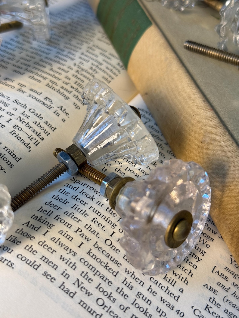 May include: Clear glass cabinet knobs with faceted detailing and brass hardware are displayed on the pages of an open book. The knobs have a vintage aesthetic, suggesting a focus on home decor and restoration projects.