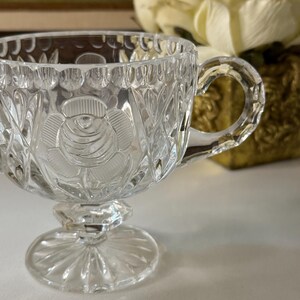May include: Clear glass teacup with a rose design on the front. The teacup has a handle and sits on a pedestal base. The glass is intricately cut with a floral and leaf pattern.