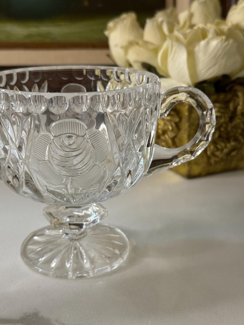 May include: Clear glass teacup with a rose design on the front. The teacup has a handle and sits on a pedestal base. The glass is intricately cut with a floral and leaf pattern.