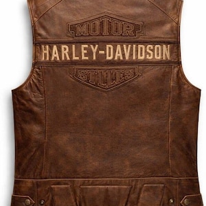 May include: Brown leather vest with the Harley-Davidson logo embroidered on the back. The vest features a classic design with a tailored fit, and two pockets at the bottom. The vest is made of a distressed leather material.