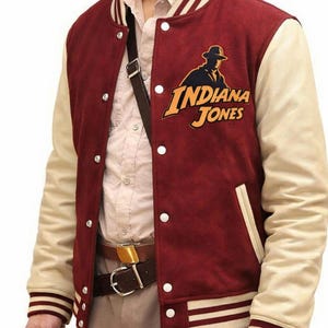 May include: A burgundy and cream varsity jacket with the Indiana Jones logo. The jacket has cream sleeves, white snap buttons, and striped cuffs. A brown leather belt and a whip are also visible.
