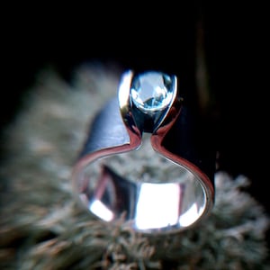 May include: A silver ring with a light blue gemstone set in a silver band. The ring is on a fuzzy, brown surface.