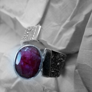 May include: A silver ring with a large, oval-shaped, deep red gemstone set in the center. The ring has a textured band.