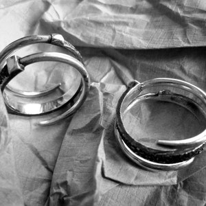 Puede incluir: Dos anillos de plata, uno con un diseño simple y el otro con un diseño más intrincado, se muestran sobre un trozo de tela arrugada.