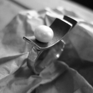 May include: A silver ring with a unique design, featuring a curved rectangular element and a white, spherical pearl. The ring is set against a textured, crumpled paper background, creating a contrast in textures.