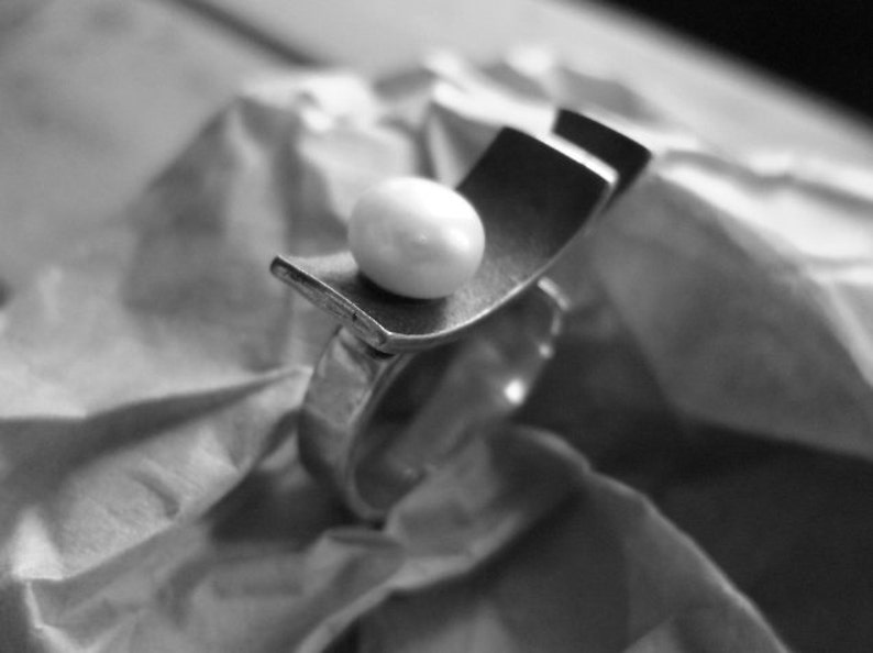 May include: A silver ring with a unique design, featuring a curved rectangular element and a white, spherical pearl. The ring is set against a textured, crumpled paper background, creating a contrast in textures.