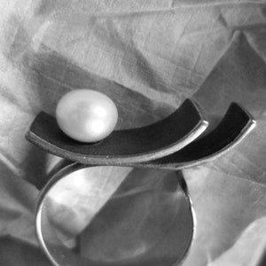 May include: A silver ring with a large, lustrous pearl set atop a dark, curved metal design. The ring's band is a simple, polished silver, creating a striking contrast with the pearl and the dark metal accent. The design is modern and elegant.