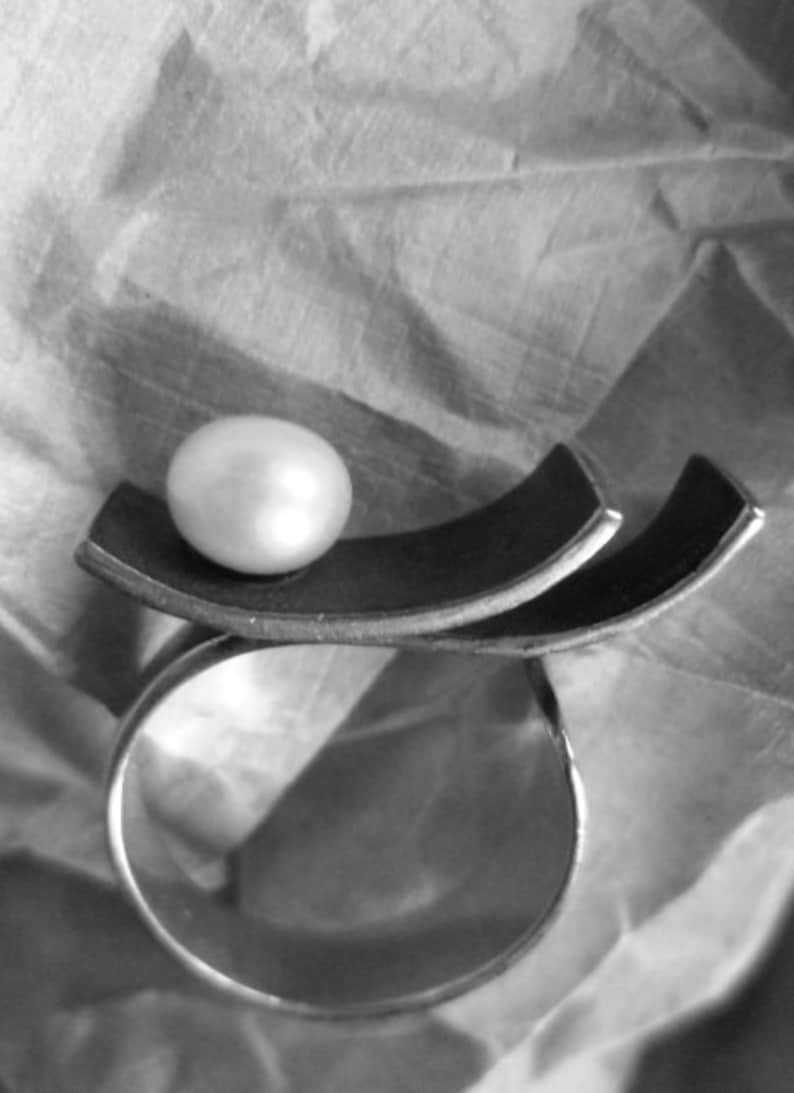 May include: A silver ring with a large, lustrous pearl set atop a dark, curved metal design. The ring's band is a simple, polished silver, creating a striking contrast with the pearl and the dark metal accent. The design is modern and elegant.