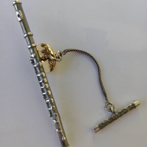 Puede incluir: Un broche de corbata de flauta de plata con una cadena y una barra de plata. La flauta tiene un acabado plateado y negro.