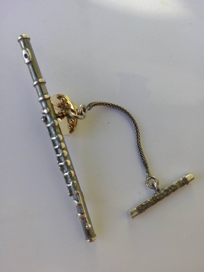 Puede incluir: Un broche de corbata de flauta de plata con una cadena y una barra de plata. La flauta tiene un acabado plateado y negro.