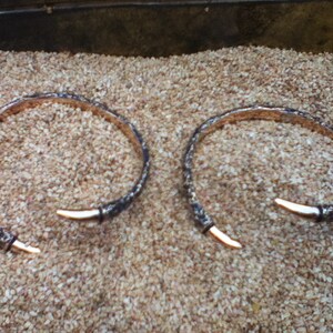 May include: Two silver bracelets with a textured finish and a pointed end. The bracelets are lying on a bed of small, brown pebbles.