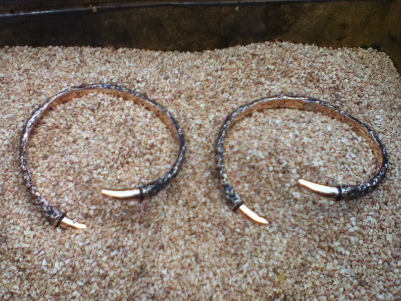 May include: Two silver bracelets with a textured finish and a pointed end. The bracelets are lying on a bed of small, brown pebbles.