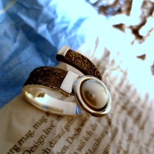 May include: A silver ring with a textured band and a round, pearl-like stone set in a silver bezel. The ring is on a white surface with text.