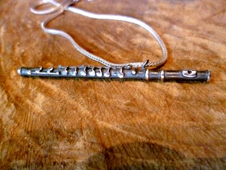 Puede incluir: Un collar de plata con forma de flauta y una cadena de plata. La flauta tiene notas musicales grabadas.