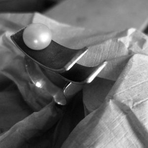 May include: A black and white image of a silver-toned ring with a large, round, white pearl. The ring has a unique, curved design. The pearl sits atop a curved section of the ring. The ring is resting on a textured, light-colored surface.