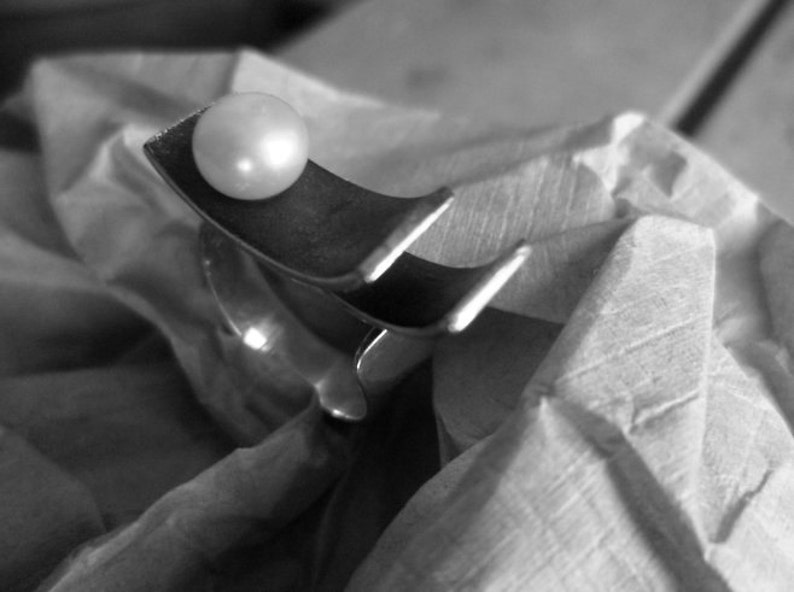 May include: A black and white image of a silver-toned ring with a large, round, white pearl. The ring has a unique, curved design. The pearl sits atop a curved section of the ring. The ring is resting on a textured, light-colored surface.