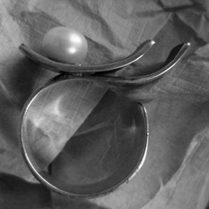 May include: A silver-toned ring with a large, round, off-white pearl. The ring has a unique design with curved, open bands. The ring is set against a textured, gray background, creating a contrast between the smooth pearl and the metal.
