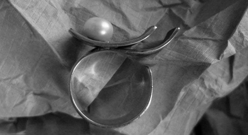 May include: A silver-toned ring with a large, round, off-white pearl. The ring has a unique design with curved, open bands. The ring is set against a textured, gray background, creating a contrast between the smooth pearl and the metal.