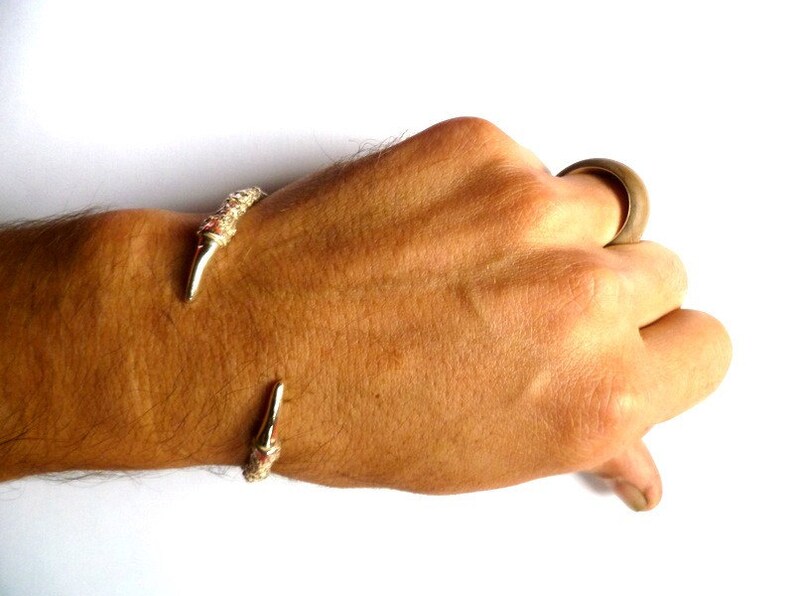 May include: A gold-toned bracelet with a claw-like design. The bracelet is worn on a person's wrist.