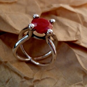 May include: A silver ring with a red gemstone set in a four-prong setting. The ring has a unique, twisted band.