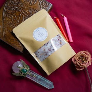 May include: A brown paper bag filled with pink bath salts and rose petals. The bag has a white label that reads "The Blessed  Botany  Salt Love  Bath Salts". The bag is on a red surface with two pink and red candles and a dried rose.