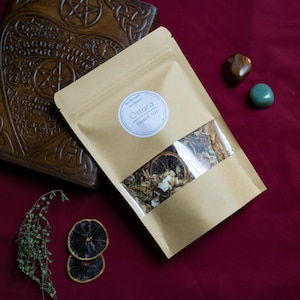 Puede incluir: Una bolsa de papel marrón con una ventana transparente que muestra hierbas secas y rodajas de naranja. La etiqueta blanca dice "Ostara Simmer Pot". Un libro de madera y dos piedras preciosas están sobre una superficie burdeos.