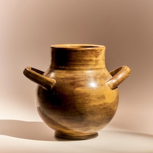 May include: A handcrafted, ceramic vase with a rounded body and two loop handles. The vase has a wide opening and a short, cylindrical neck. The color is a blend of brown and tan, with darker streaks throughout. The vase sits on a small, circular base.
