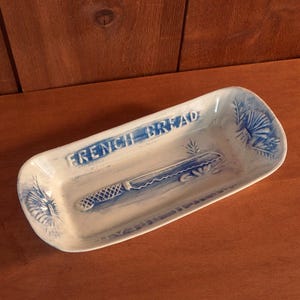 Può includere: Un piatto da pane rettangolare in ceramica con un design blu e bianco. Le parole "FRENCH BREAD" sono scritte in blu in alto. Un disegno di coltello è al centro e motivi floreali sui lati.