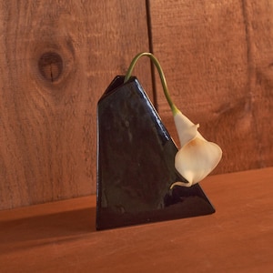 May include: A black triangular ceramic vase holding a single white calla lily. The vase has a glossy finish and is set against a wooden background. The flower's stem curves gracefully.