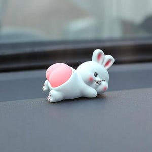 May include: A small, white bunny figurine with a pink bottom, lying on its stomach with a winking expression. The bunny has pink inner ears and is set against a dark gray surface.