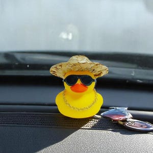 May include: A yellow rubber duck wearing a straw hat, black sunglasses, and a gold chain necklace. The duck is positioned on a car dashboard, with a blurred background of a windshield and sky.