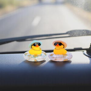 Op de afbeelding: Twee gele badeenden met zonnebrillen, hoeden en gouden kettingen, elk in een doorzichtige opblaasbare ring. De eenden staan op het dashboard van een auto, met een weg en een voorruit op de achtergrond.