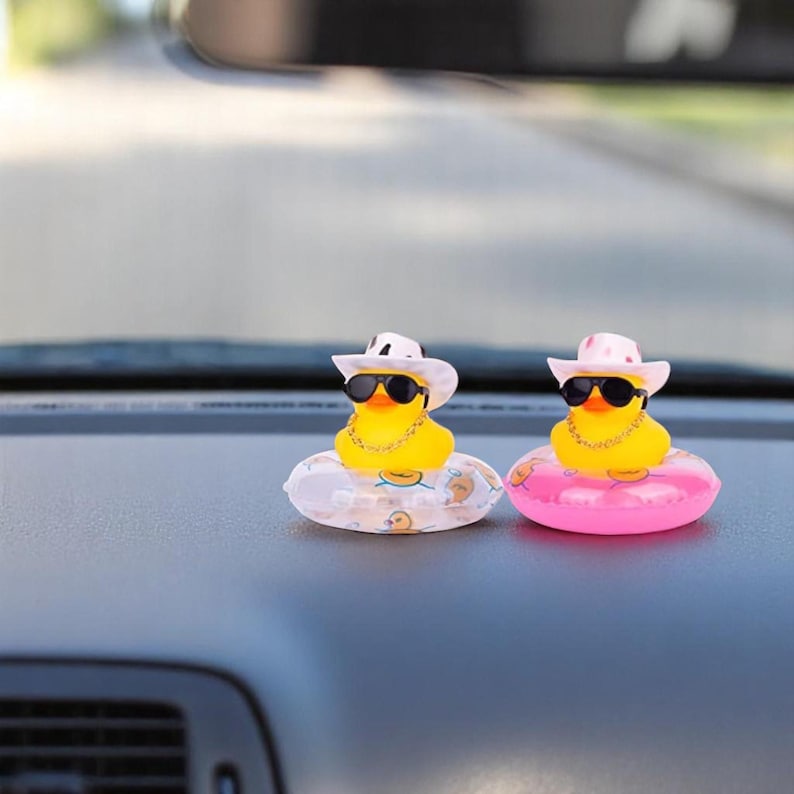 Op de afbeelding: Twee gele badeenden met zonnebrillen, cowboyhoeden en gouden kettingen, elk drijvend op een kleine opblaasbare ring. E&eacute;n ring is bedrukt, de andere is roze. De eenden staan op een autodashboard.