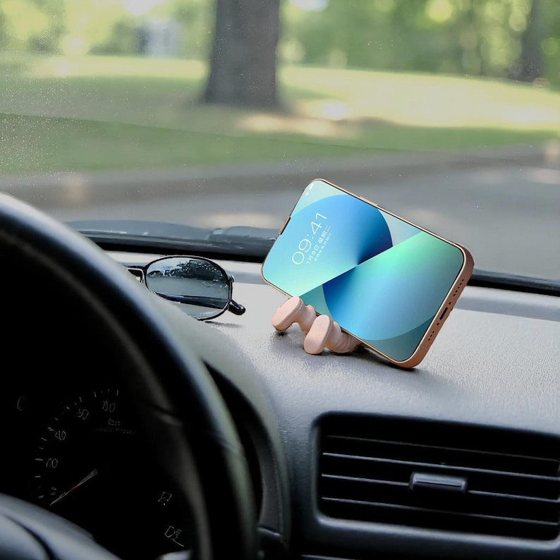 Op de afbeelding: Een smartphone met een goudkleurige behuizing rust in een lichtgekleurde, houten telefoonhouder op het dashboard van een auto. Een zwarte zonnebril ligt naast de telefoon. Het telefoonscherm toont een blauw-groene gradi&euml;nt.