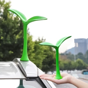 May include: Two green plastic sprout-shaped car antenna toppers. The larger topper is on top of a car, while the smaller topper is held in a person's hand.