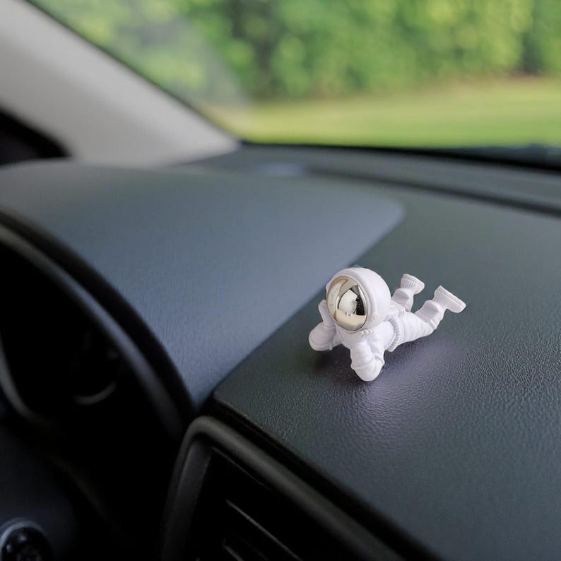 Op de afbeelding: Een witte astronautenfiguur met een zilveren helm ligt op een donkergrijs dashboard. De miniatuur astronaut ligt in een liggende positie, wat een speelse toets geeft aan het interieur van de auto. De achtergrond is wazig, wat duidt op een buitenomgeving.