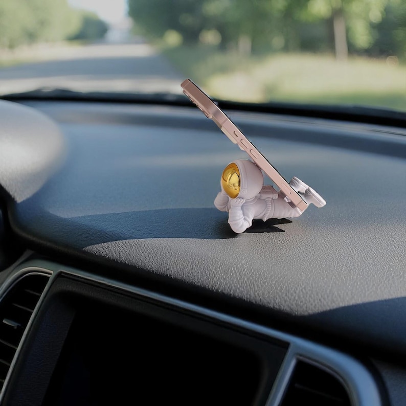 Op de afbeelding: Een witte astronaut-vormige telefoonhouder met een goudkleurige helm ondersteunt een ros&eacute;gouden telefoon. De telefoonhouder staat op het dashboard van een auto, met een vage achtergrond van een weg en bomen.