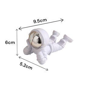 Op de afbeelding: Een witte astronautenfiguur, liggend, met een reflecterend gouden vizier. De figuur is 9,5 cm lang, 6 cm hoog en 5,2 cm breed. De astronaut bevindt zich in een ontspannen houding.