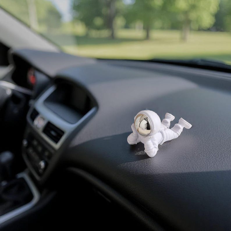 Op de afbeelding: Een witte astronautenfiguur met een reflecterende zilveren helm ligt op het dashboard van een auto. De figuur bevindt zich in een ontspannen pose, met uitgestrekte armen en benen. Het interieur van de auto is zwart en grijs.